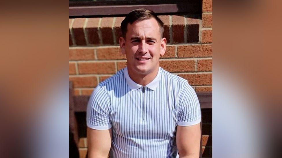 A man in a white and blue stripe zip up T-shirt looks at the camera.