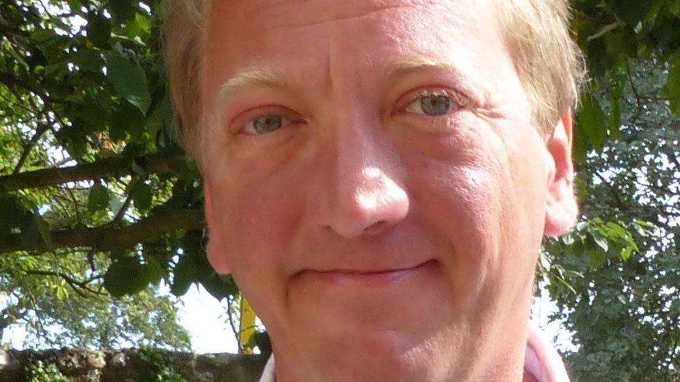 A close-up of Adrian Marsden, looking towards the camera and slightly smiling. He has fair, short hair and is clean-shaven. Behind him is a canopy of green leaves from trees and through them glimpses of sky 