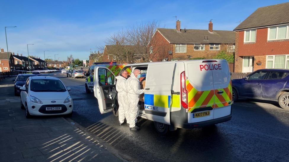 A police van parked in a residential street, with two men, wearing white, 'haz-mat' overalls, standing alongside.