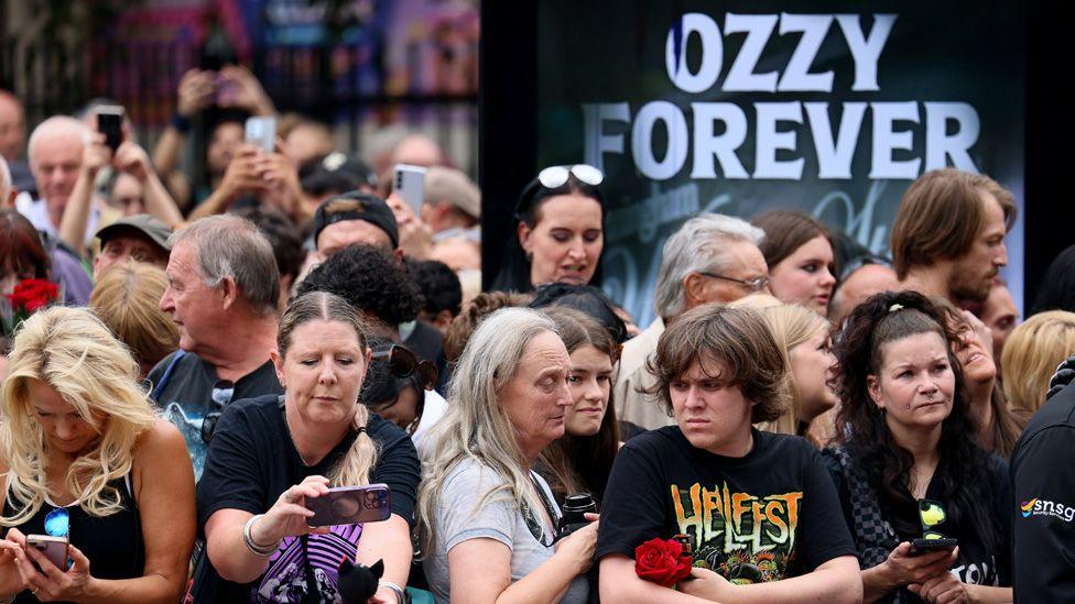 A crowd of Ozzy fans standing in front of an advertising screen showing the words "Ozzy forever"