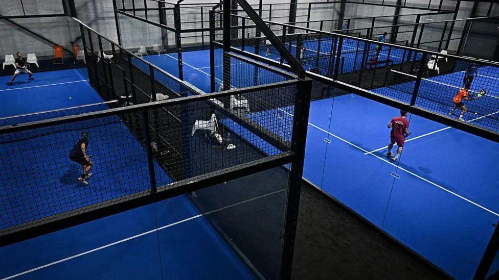 An indoor padel court centre, with blue courts. A number of people are engaged in matches, with each court separated by black metal fences, nets and glass.