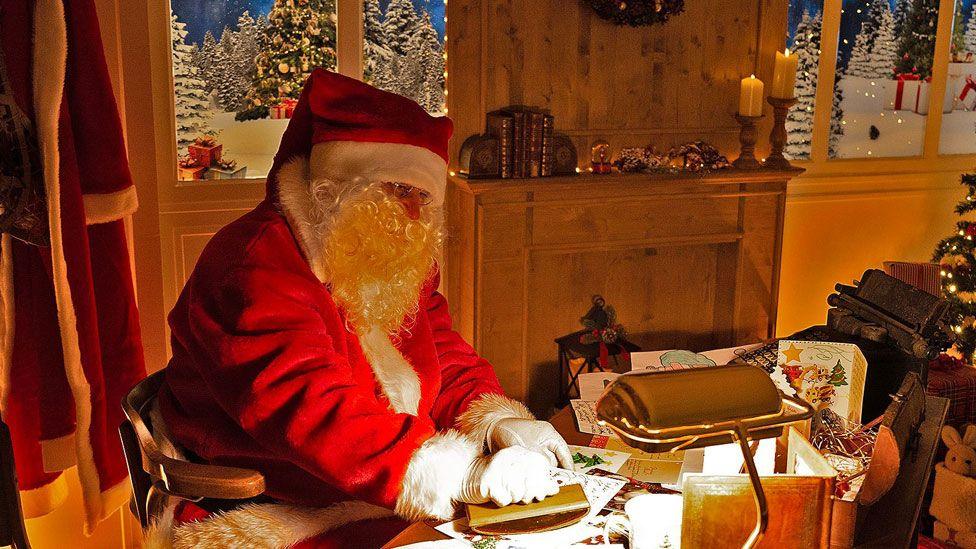 A man dressed as Father Christmas in a red suit trimmed with white fur and wearing a white-trimmed fur hood. He is sitting at a desk which is scattered with letters. He has a long white beard and is wearing glasses. He is sitting in a room with a fire place and a Christmas tree can just be glimpsed on his right. Image from Libourne, France, in 2023