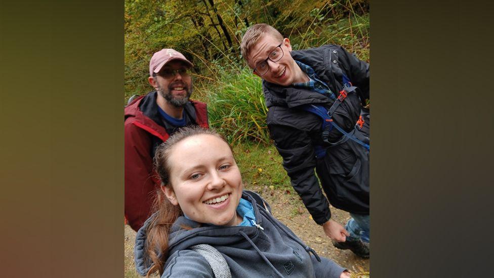 Hannah taking a selfie along with two male friends on a walk through woodland - the angle is slightly sideways. She wears a grey hoodie and has her hair in a ponytail. One man wears glasses and has a black coat with a hiking rucksack, and the other has a red coat and baseball cap, and a grey beard. 