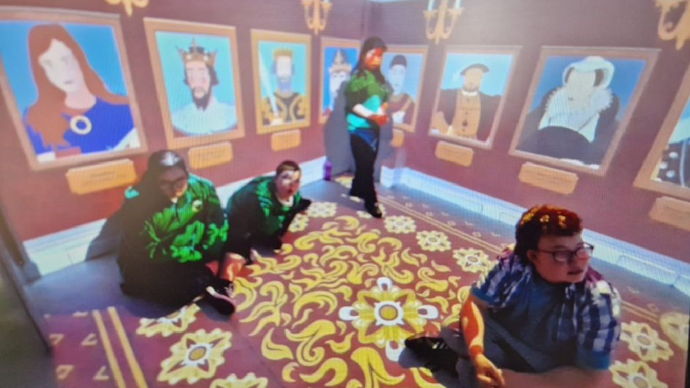 A picture of the immersive room being used in an educational lesson at Greenfield school. There are three students and a teacher inside. The two walls have portraits of kings and queens on. The floor is also lit up with yellow flowers and various moving patterns.