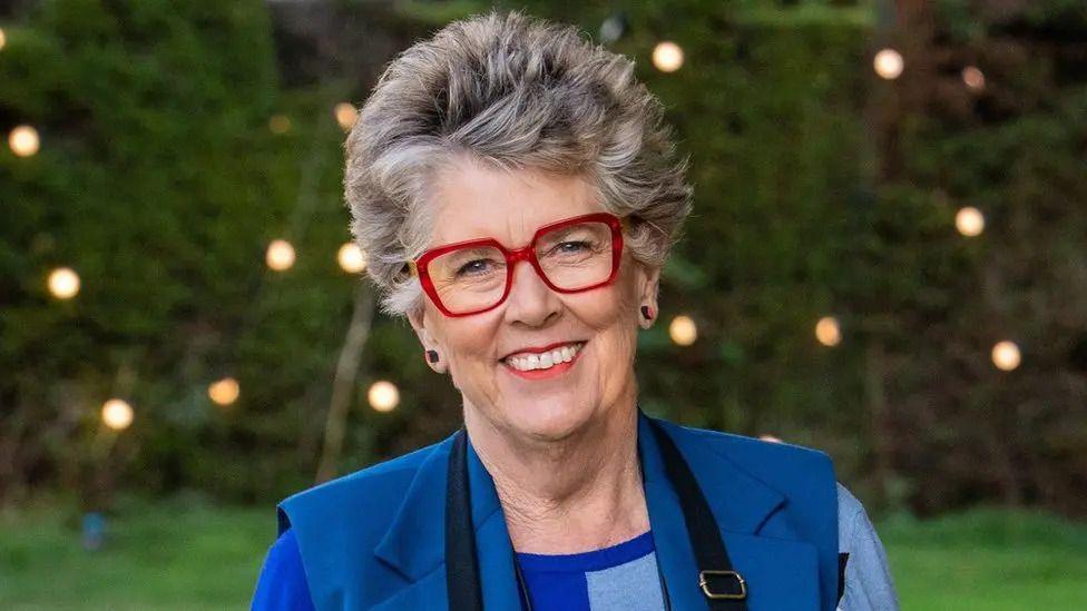 Dame Prue smiling, wearing a blue outfit and red glasses