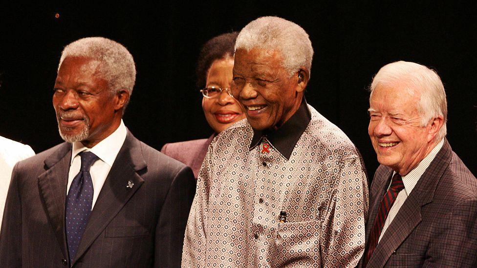 Jimmy Carter, wearing a brown jacket, white shirt and red tie, is standing on the right of a line-up of three former global leaders. In the centre is Nelson Mandela who is wearing a silver-coloured shirt with a black collar. On the far left is former UN Secretary-General Kofi Annan in a black jacket, white shirt and blue tie. All three men are smiling as if they are posing for a picture.