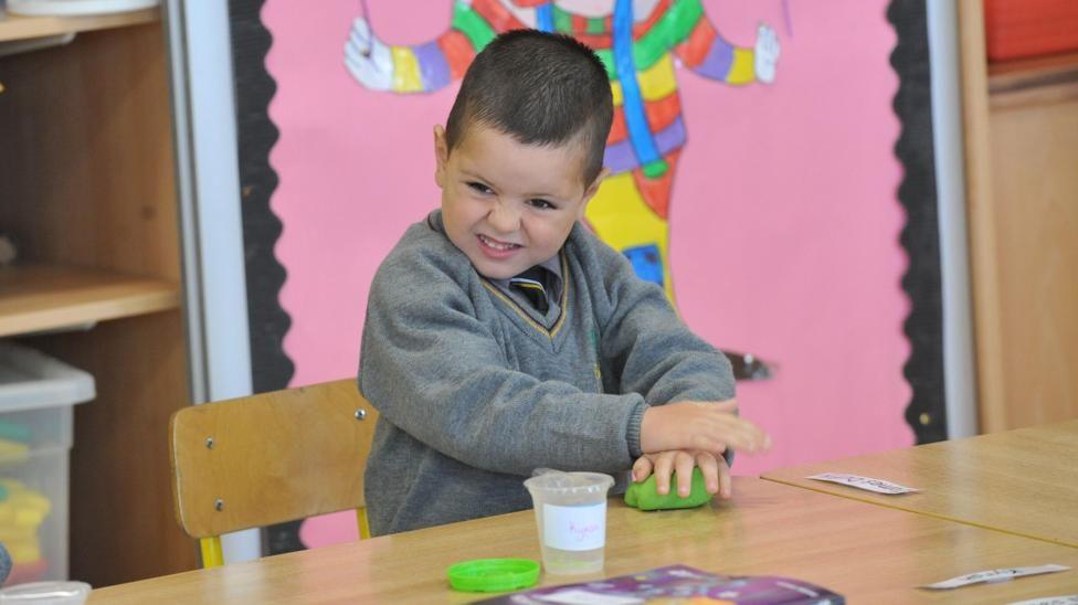 Kyran Durnin pulls a face while playing with green play dough in a primary classroomera. He has short brown hair and brown eyes. Kyran is stood over a wooden table, with a small wooden chair behind him, a pink colourful display is on wall behind him He is wearing a grey jumper with black tie