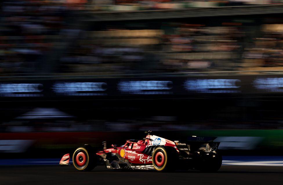 A red Formula 1 car with Ferrari branding speeds along a racetrack, captured in motion blur against the backdrop of a grandstand, emphasizing its sleek design and high-speed action.