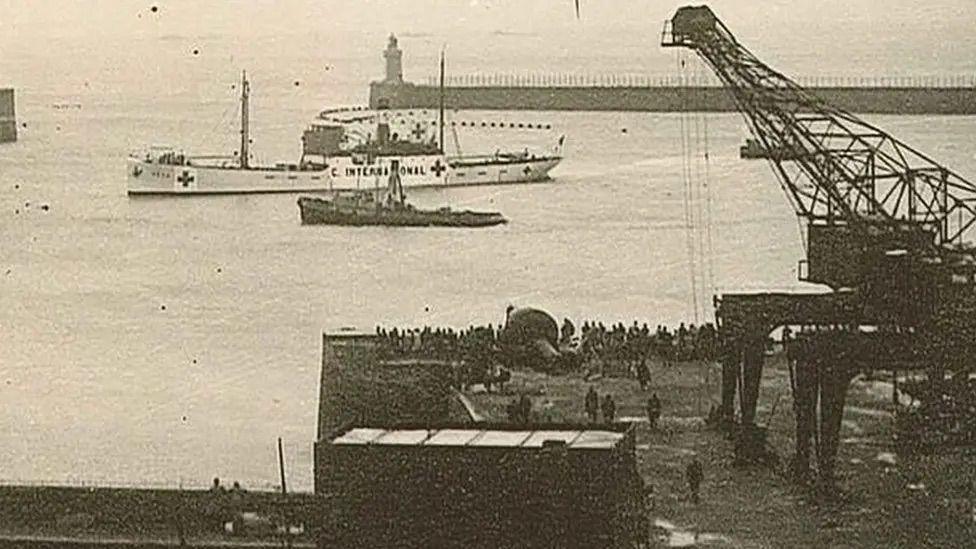 A black and white image of The SS Vega ship in the water in Guernsey's Saint Peter Port harbour in 1944. There is a tug boat nearby and hundreds of people are waiting on the harbour net to a large crane.