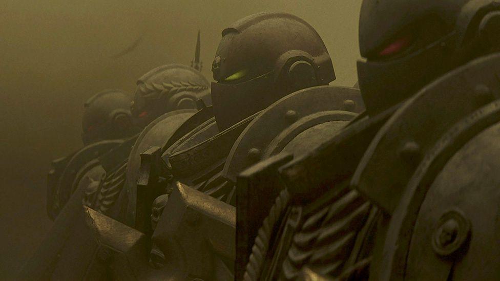 Computer generated image shows a line of four heavily armoured troopers staring at an unseen foe. Their gunmetal grey suits of armour are metallic and industrial looking, with bolts and rivets visible. The group is hard to distinguish from the dark grey dust swirling around them.