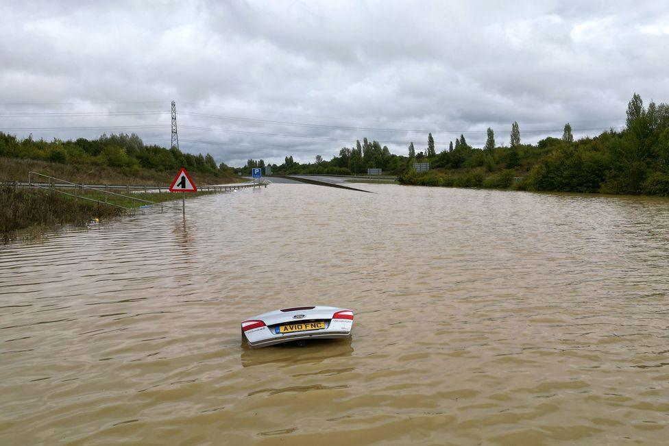 The open boot of a car is visible above the water where the vehicle is submerged in flood water on A421 in Marston Moretaine, Bedfordshire.