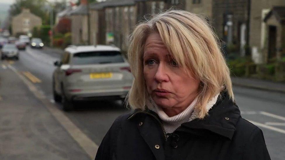 A woman with blonde hair and wearing a cream jumper and black coat stands on the pavement next to a road. In the background a grey car is driving away from her and a car on the other side of the road has its headlights on. 