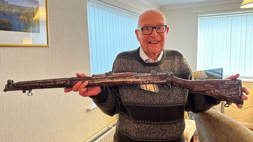 Roy Hayward wearing a striped greeny-brown jumper over a white shirt  holding up a Lee-Enfield Rifle, standing in a sitting room with windows and yellow chairs behind him