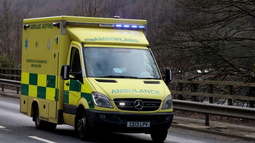 A ambulance on an emergency call in Abercarn, Wales. The vehicle can be seen on the road with metal barriers behind it, and tree branches in the background. 