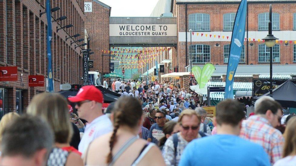 A crowd of people walking around Gloucester Quays. So0me in the foreground are out of focus. There is a sign in the distances on a footway bridge that says 'Gloucester Quays'