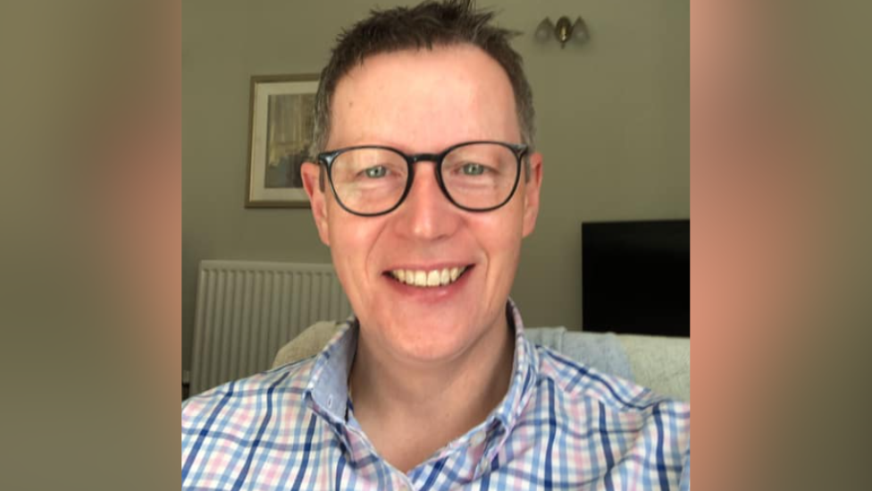 A man in a blue and pink and white checked shirt, wearing black glasses and short grey hair