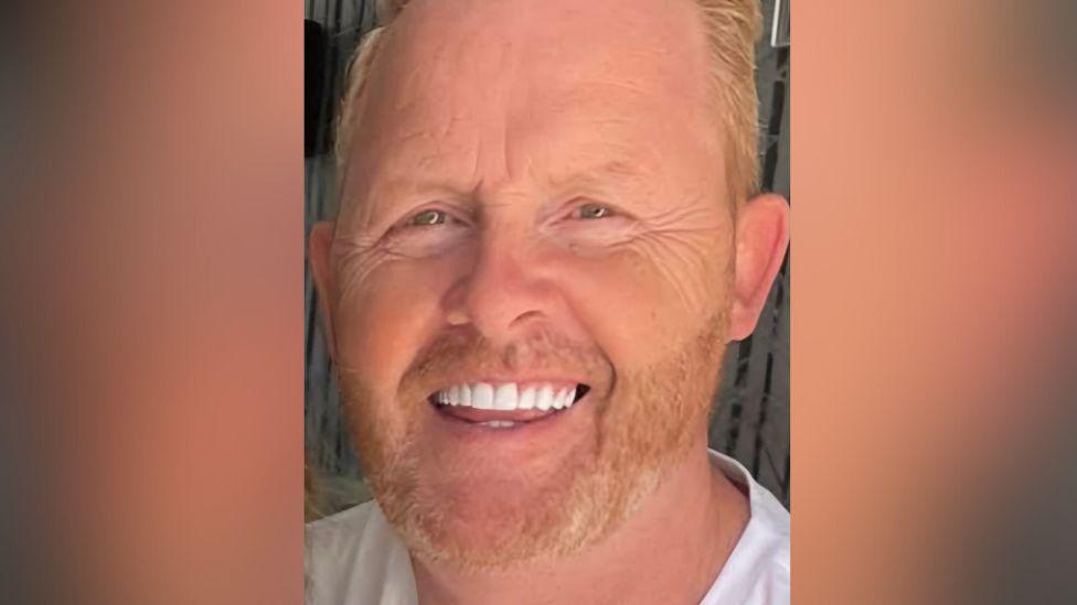 Dennis O'Leary, middle-aged man with fair hair, stubble and white teeth. He is smiling at the camera.