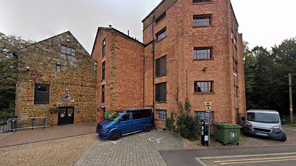 The outside of Towcester Mill Brewery, showing a couple of vans parked outside a large ironstone building with a big brick extension. Above the main entrance is a union flag.