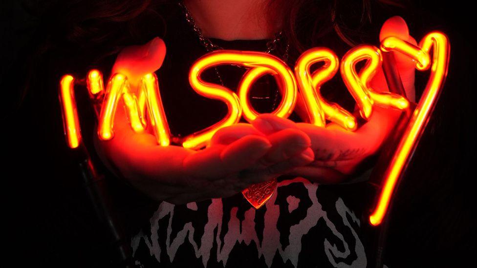 One of the portraits by artist Annabel McCourt showing a close up of a pair of hands holding an orange neon sign saying I'm Sorry