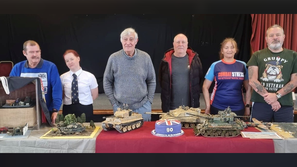 A group of people standing in front of a table with a numerous items on it. There are models of tanks and a blue, red and white cake with the number 80 on it.