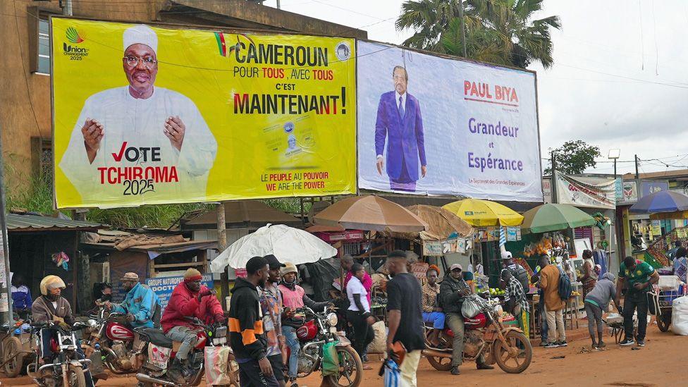 People hang around on motorbikes on a dustry road where two campaign posters have been erected - one for Biya and the other for Tchiroma.