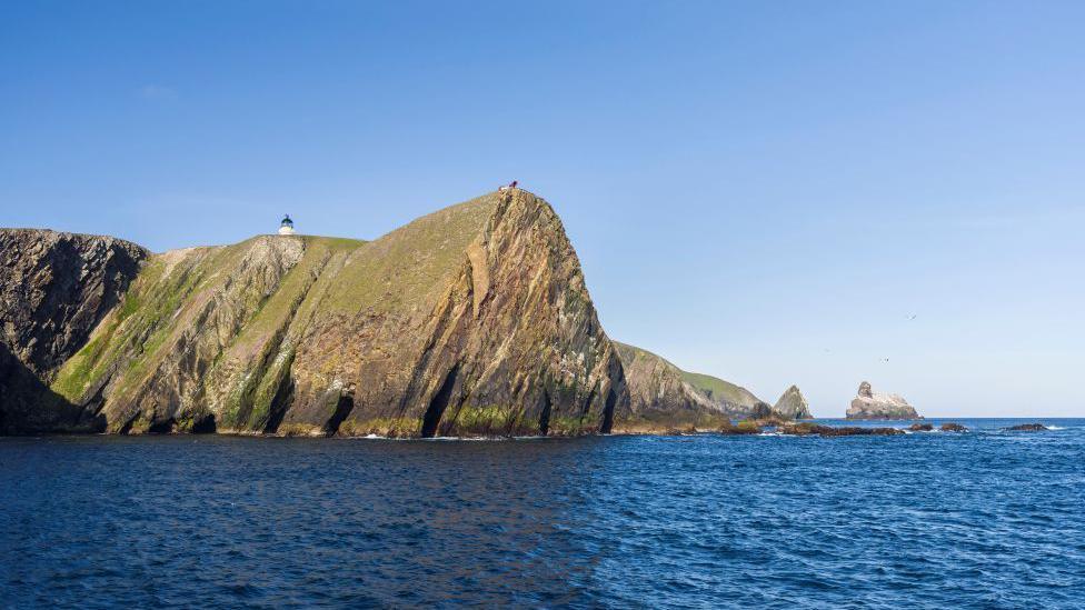 Image of Fair Isle taken from the sea, blue water and a blue sky, with the top of a lighthouse just visible.