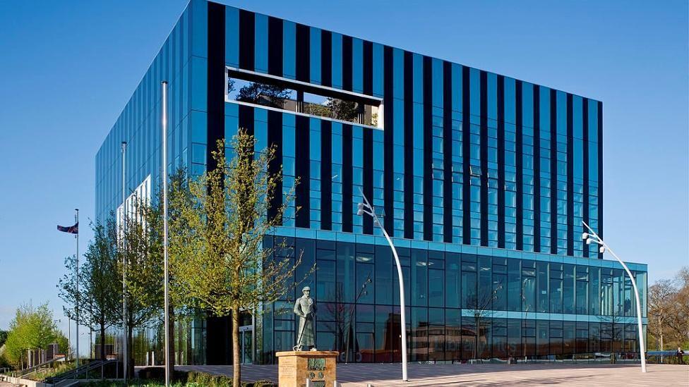 Large steel and glass building with tall windows on the ground floor and trees to one side. There is a statue on a plinth in front of the building.