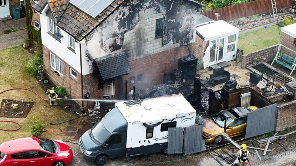 Two firefighters using hoses on a gold car, camper van and smouldering shed remains