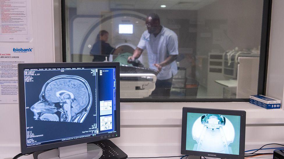 A shot of the operation room at UK Biobank. In the distance is a window showing a person being scanned in an MRI machine. They are being attended to by two radiographers operating the machine. In the foreground is a picture of a brain scan on a monitor and another video screen showing the internal MRI scanner tube.