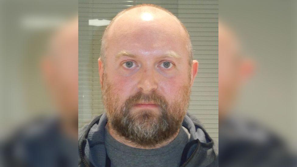 Police custody photograph of Karl Abbott who is bald with a dark beard and wearing a grey T-Shirt and jacket