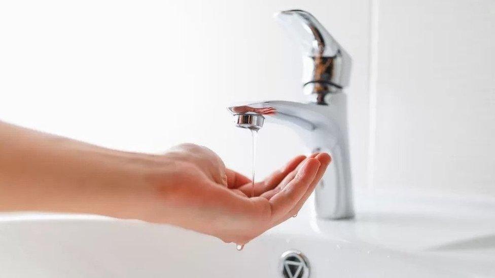 A silver tape with low water pressure. A hand is collecting the water and there are white tiles in the background.