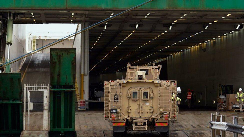 Cavernous inside of cargo ship with browm armoured army vehicle driving onboard.