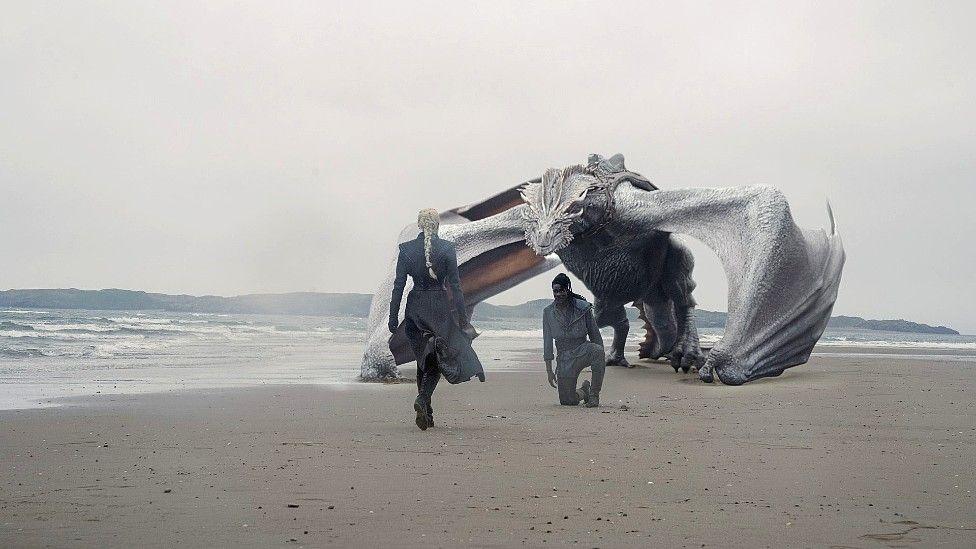 Scene from House of the Dragon - two actors on a beach, one kneeling, with a dragon behind him