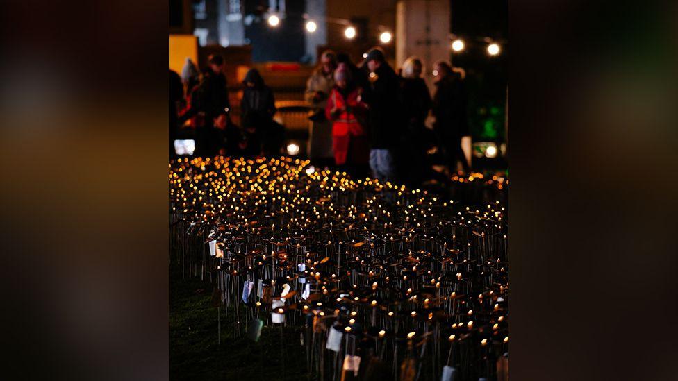 Hundreds of steel flowers lit up in the garden of St Luke's Bombed Out Church in the evening with people blurred out in the background looking at the artwork.