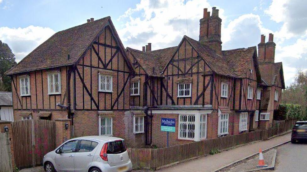 The house that was formerly the Barn School in Much Hadham. It is a large two-storey half-timbered red brick building with roofs and gables pointing in different directions. There is a For Sale sign at the front a silver car is parked on the driveway.