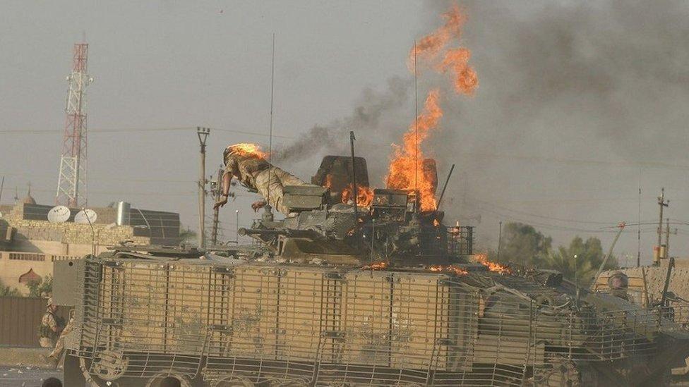 A military tank has flames coming from the top. A soldier whose clothing is on fire appears to be falling or jumping from the tank.