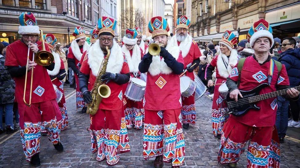 A group of men dressed in festive outfits play brass instruments and a bass guitar while walking in the parade. A row of drummers can be seen behind them as crowds look on either side. 