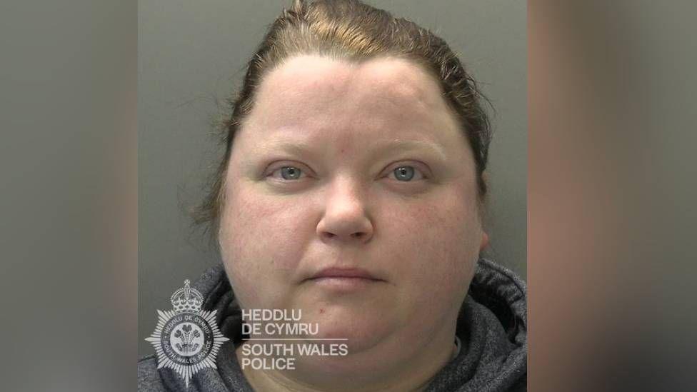 Jemma Lewis has blue eyes and brown hair. She is wearing a top with a grey collar. The top of the Gwent Police logo can be seen in this police mugshot.