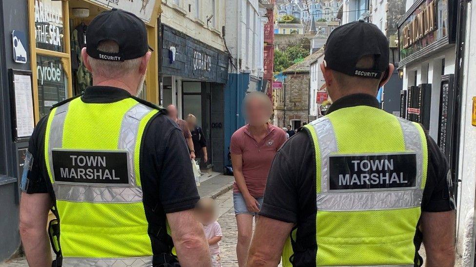 Two street marshals with their backs to the camera in a St Ives street. They both are wearing yellow hi-vis. 
