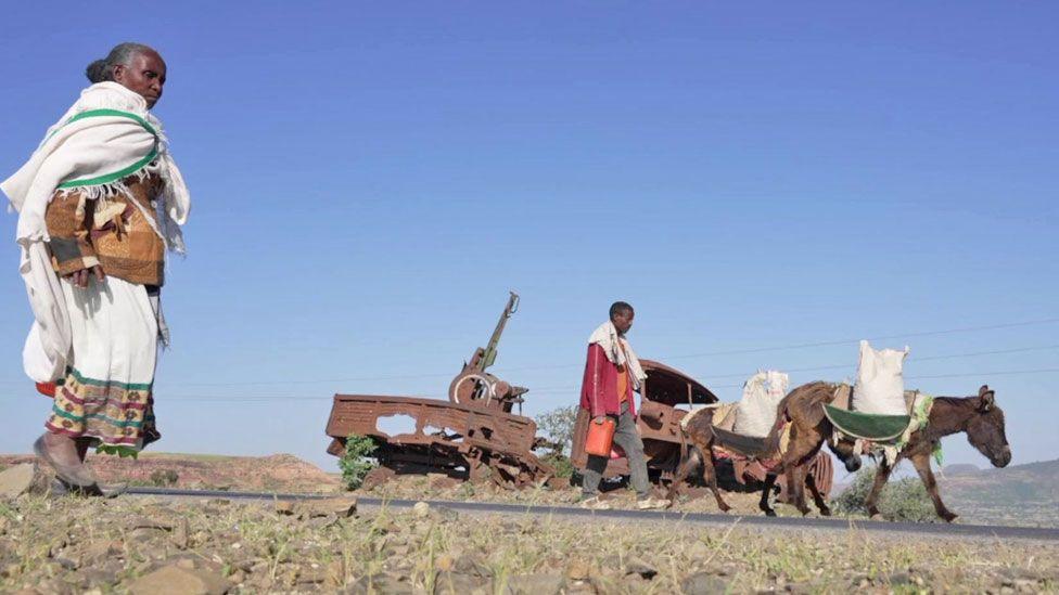 A military vehicle with a big gun rusts by the side of a main road in rural Tigray as a woman and man walk by. The man is following behind two mules, which are carrying sacks.