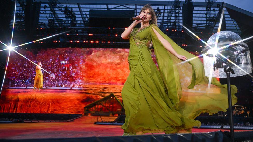 Taylor Swift performing at Anfield in a green dress in front of a big screen showing her and the crowd