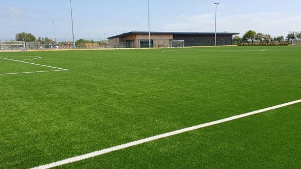 A football pitch at Common Edge Community Sports Village, showing a huge expanse of grass market out for football, with a single storey clubhouse in the background behind three floodlight poles