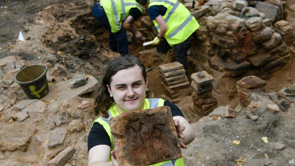 A volunteer at a dig site holding up an Imperial tile. Two workers are excavating part of the site containing an ancient wall behind them.