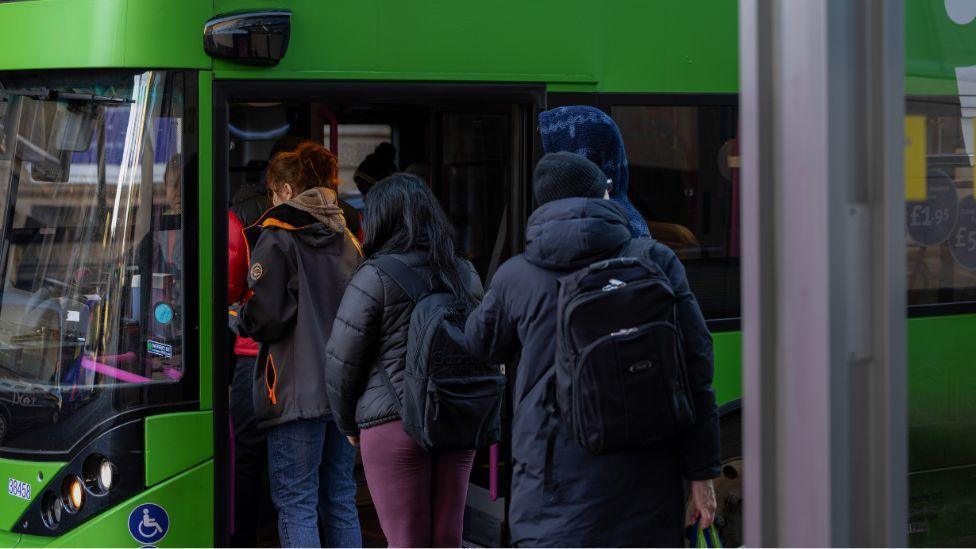 People board a bus in Glasgow. Three people, dressed in dark jackets, are awaiting in a row to get on a green coloured s