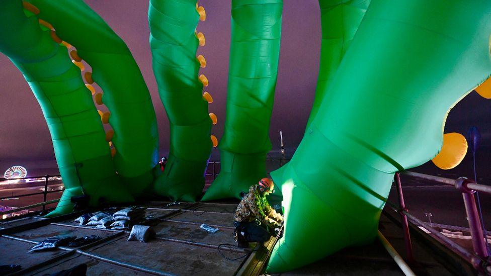 Six huge inflated tentacles being installed on a rooftop at night by a workman who is dwarfed by them. The tentacles are green with yellow nodules and they rise up into the air