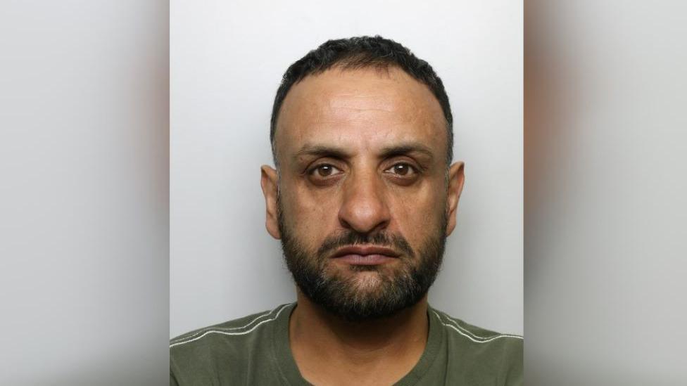 A mugshot image of Asuk Hussain, 53, from Dewsbury, where he looks solemnly at the camera, has black hair and is wearing a green top with white stripes