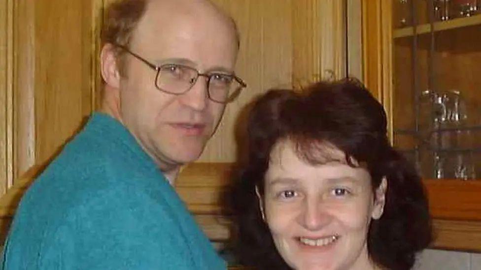 A man wearing a blue jumper and sporting spectacles stands with a woman who has brown hair, both are looking at the camera