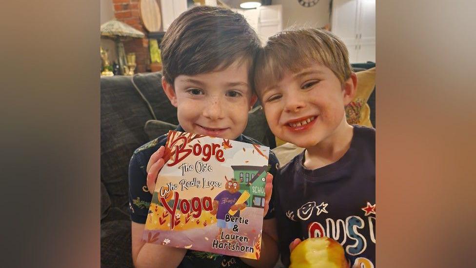 Bertie, aged seven, has brown hair and is holding a copy of Bogre The Ogre Who Really Loves Yoga. On the cover is a brown ogre in a purple top, and he is holding a yellow yoga mat. next to Bertie is his brother Ralph, who has light brown hair and is holding an apple.