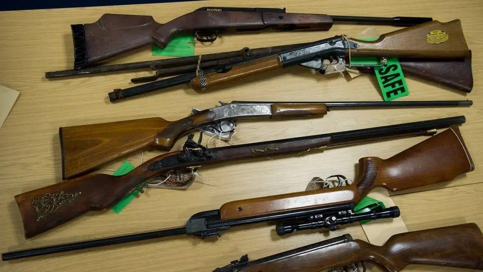 Seven brown rifles lying on a wooden table with green labels stating "SAFE" attached. Some have sights attached.