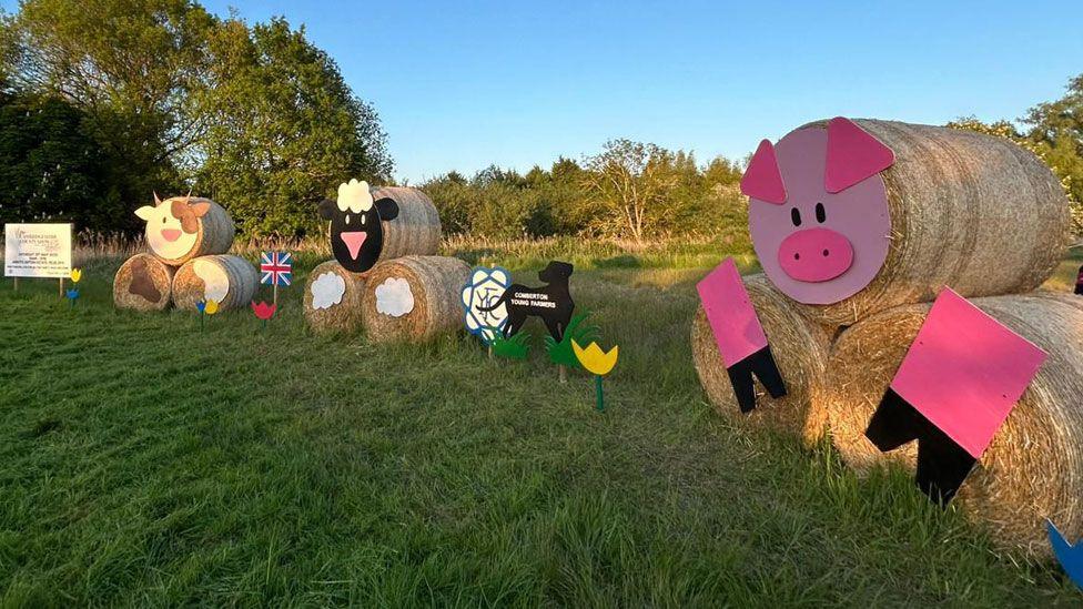 Three straw bale sculptures made from round bales, one resting on top of two at the base. On the left, one has a face of cow, in the middle is one with the face of a sheep, and the third has the face of a pig. Between two and three is a cut-out of a dog.